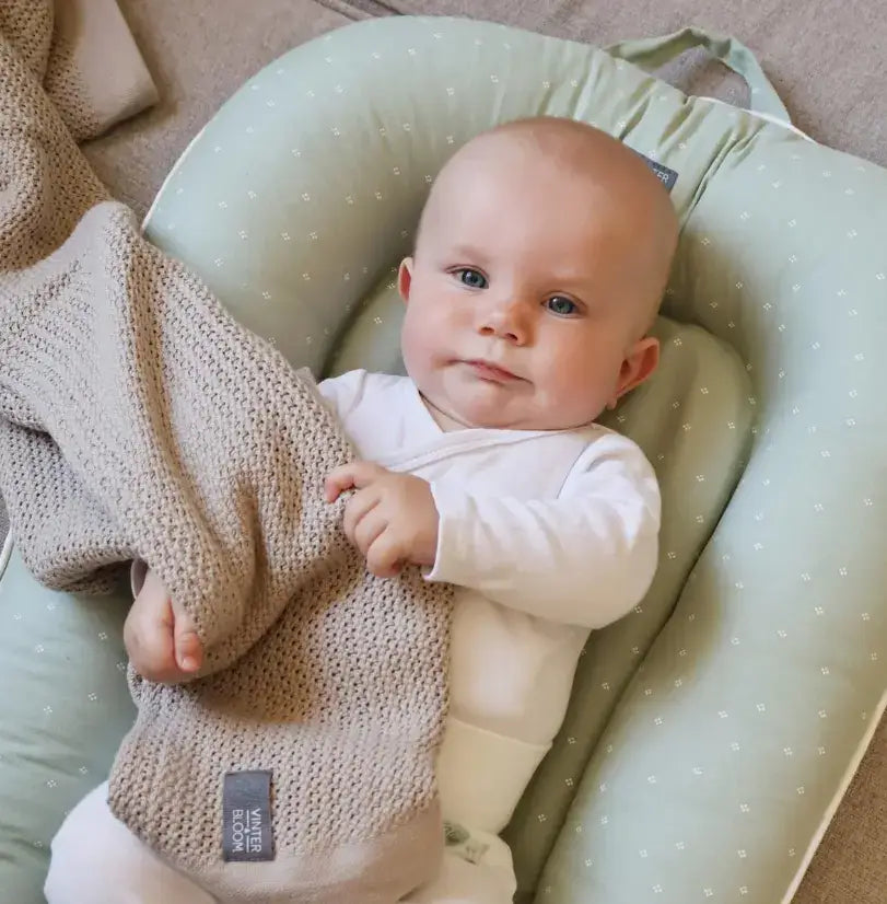 Baby ligger i Vinter & Bloom babynest med beige stickad filt.